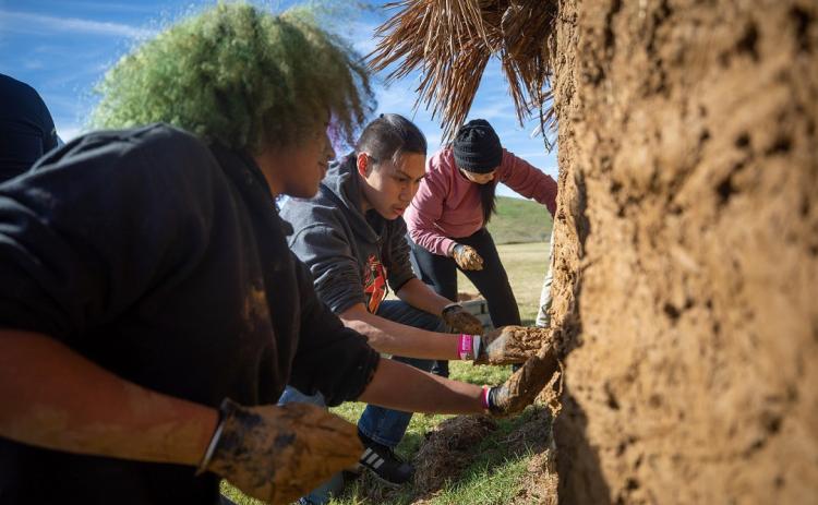 From left to right Arianna Myers, Colt, James and Cheyhoma Dugger all work together to throw and spread clay on the outside of a traditional Choctaw shelter. Photo provided | Choctaw Nation